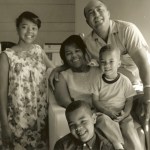 A black and white photo of a family consisting of a father, mother, two young children, and an older girl, all smiling and posing together.